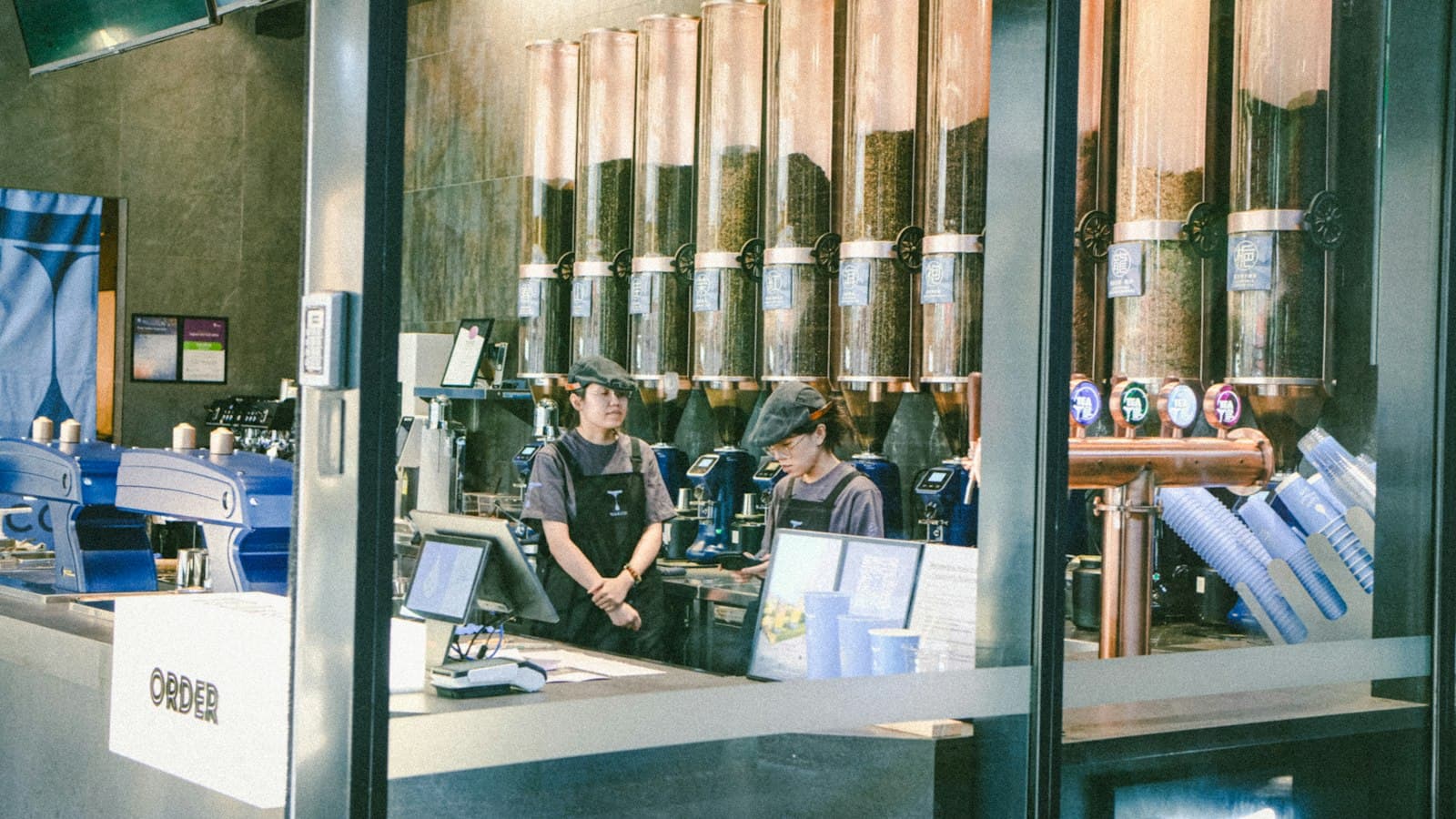 Two baristas behind a tea shop counter with a wall of tea tins